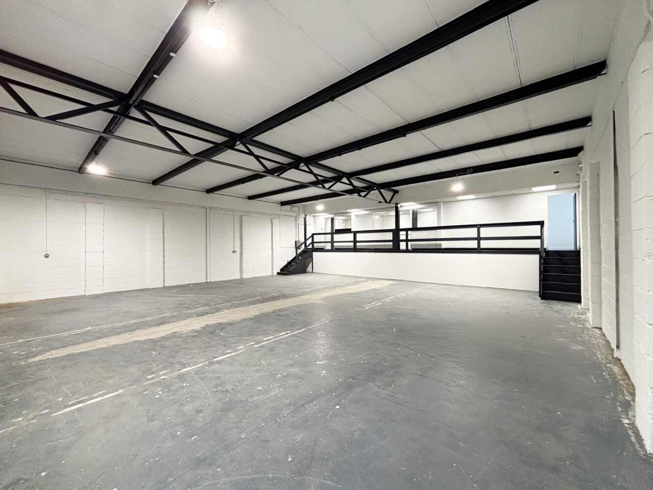 Spacious empty warehouse with white walls, exposed black ceiling beams, concrete floor, and a raised mezzanine level with black railings and two staircases.