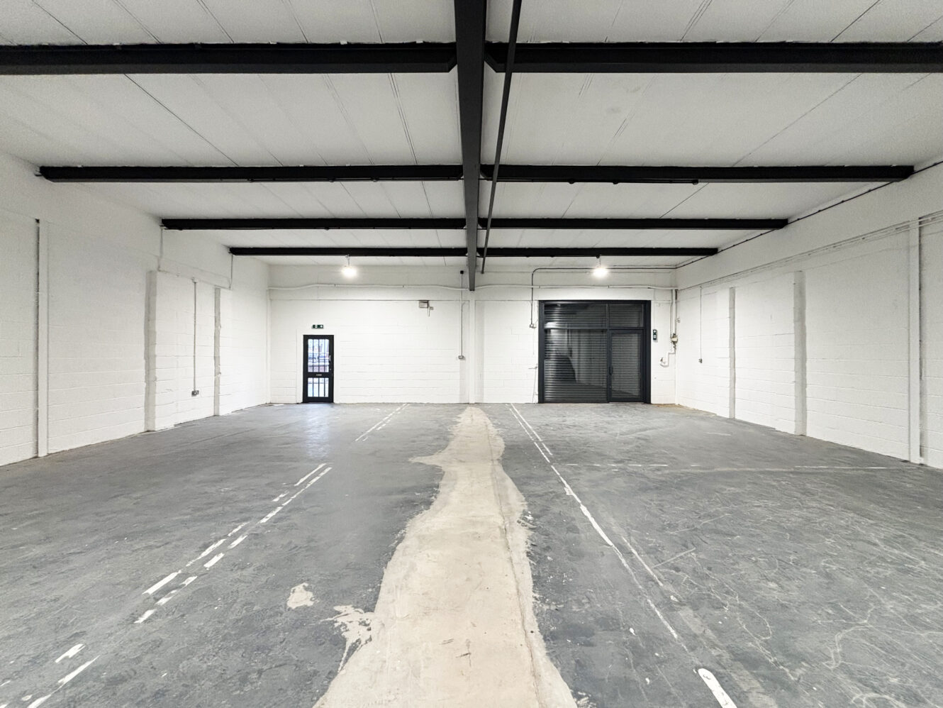 Empty industrial warehouse space with concrete floor, white walls, visible ceiling beams, a black roller shutter door, and a single side exit door.