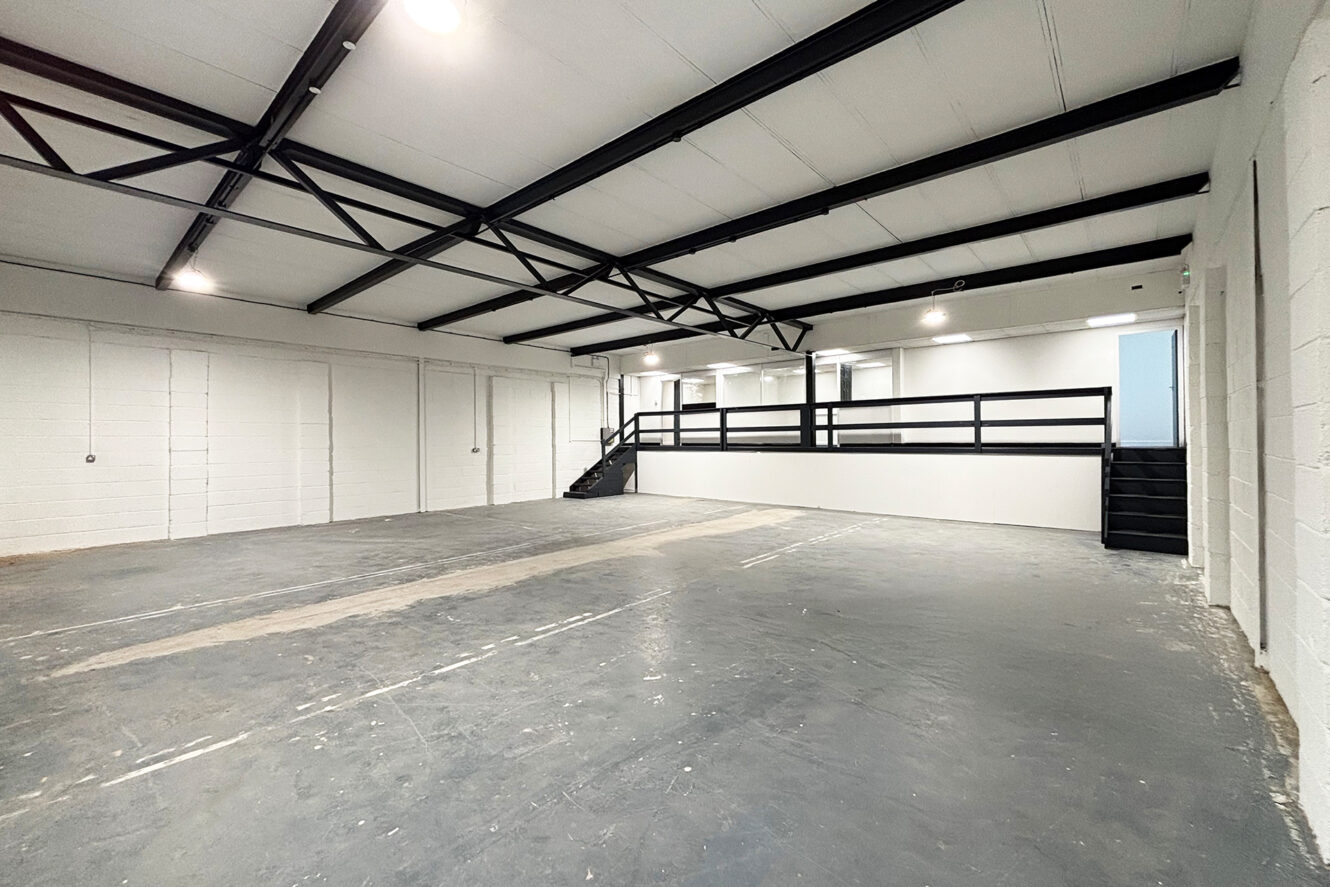 Large empty industrial space with white walls, exposed ceiling beams, concrete floor, and a mezzanine level accessible by two staircases.