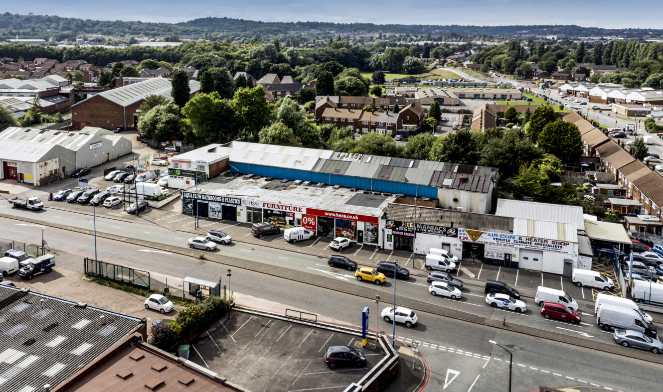 A busy street with shops and warehouses on one side, cars and vans on the road, and residential buildings and greenery in the background.