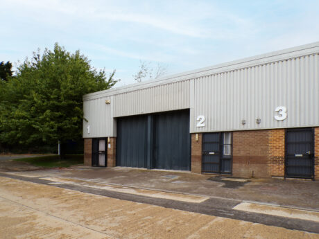 Industrial building with three numbered units (1, 2, and 3), each with a door and the middle unit (2) having a large central shutter, bordered by trees and a paved area.