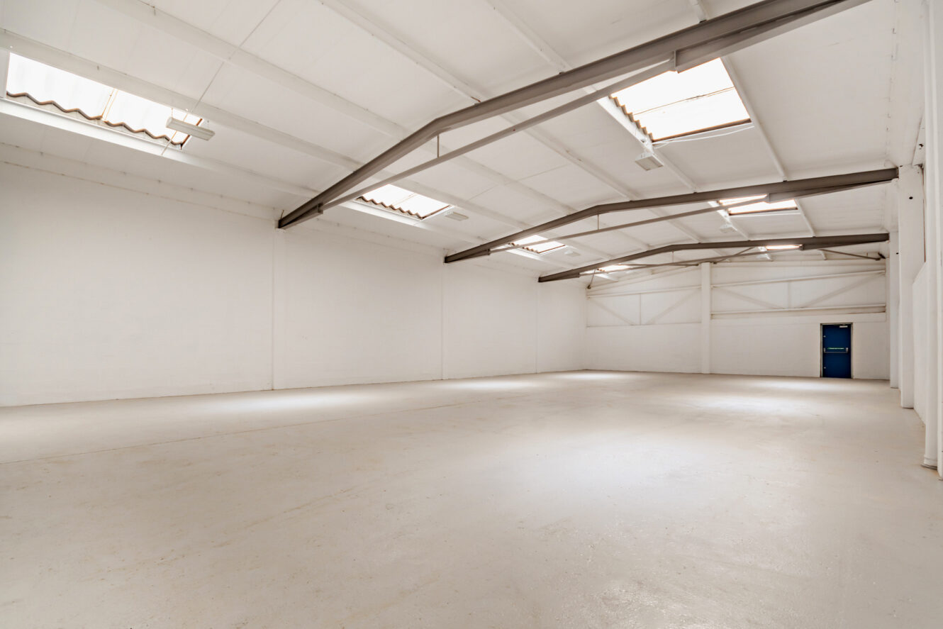 A spacious, empty warehouse with white walls, a clean floor, exposed beams, skylights, and a single blue door on the far wall.