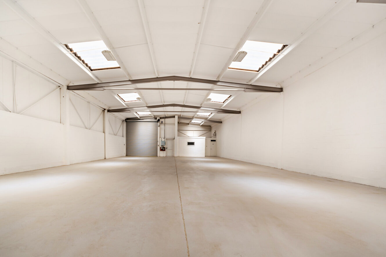 Interior of an empty warehouse with white walls, high ceiling, skylights, and a large closed roller door at the back.