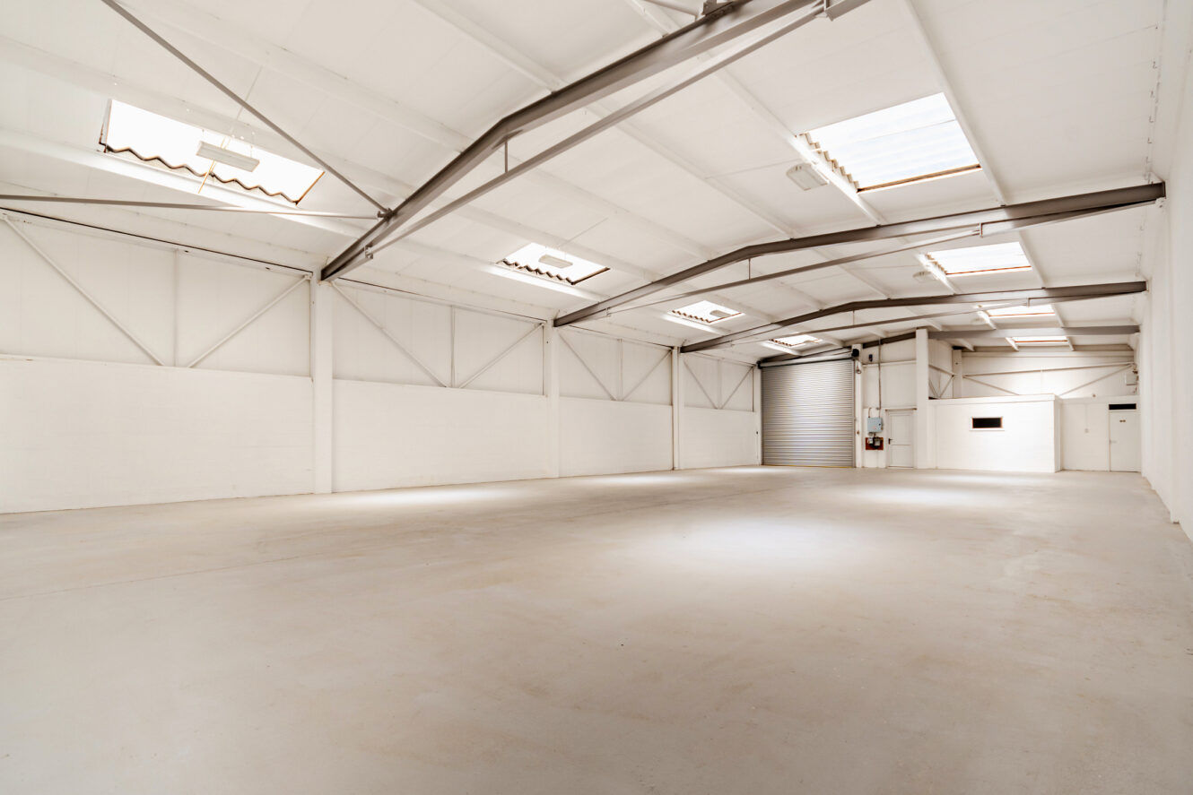 Spacious empty warehouse with white walls, high ceilings, skylights, and a large roller door.