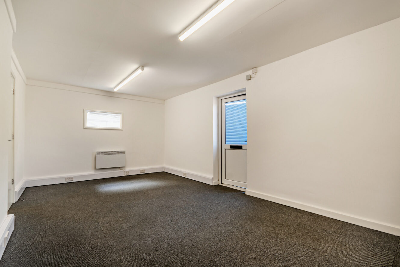 Empty room with white walls, grey carpeted floor, fluorescent ceiling lights, a small window, wall heater, and a door leading outside.