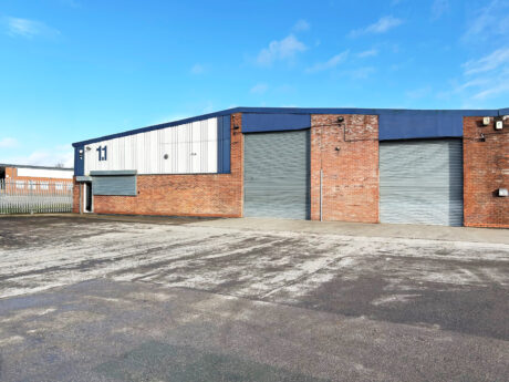 A large industrial warehouse with blue and white paneling, brick walls, three closed roller doors, and the number 11 displayed on the building. The yard in front is empty.