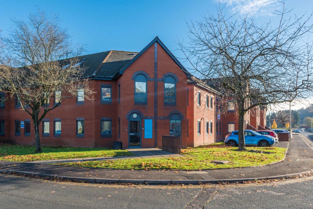 A modern two-story red brick office building with arched windows, surrounded by bare trees, parked cars, and a clear blue sky.