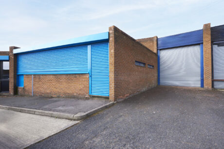 A brick industrial building with two blue metal roller shutter doors and a concrete driveway in front.