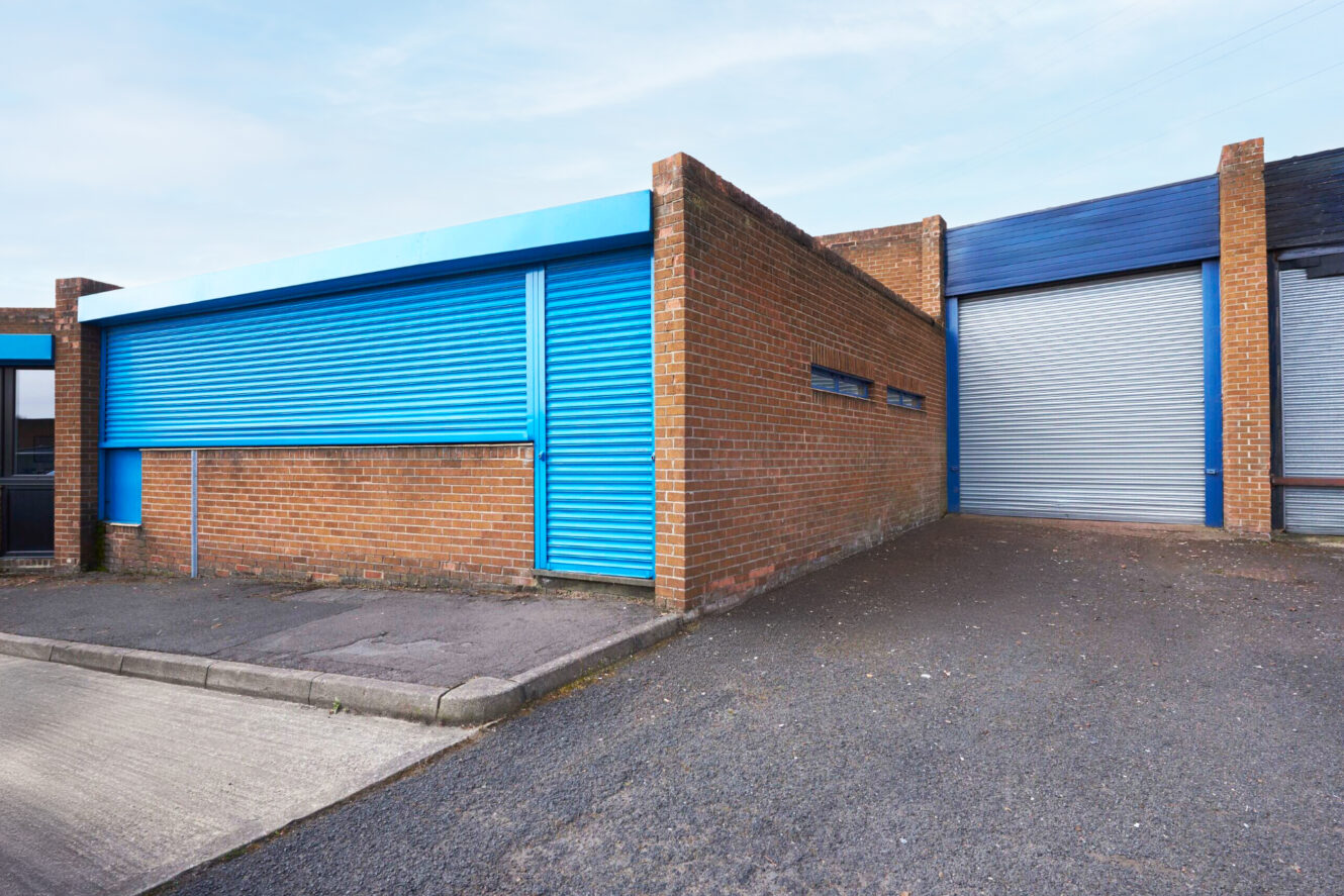 A brick industrial building with blue metal roller shutter doors and small horizontal windows, set on an asphalt and concrete lot.