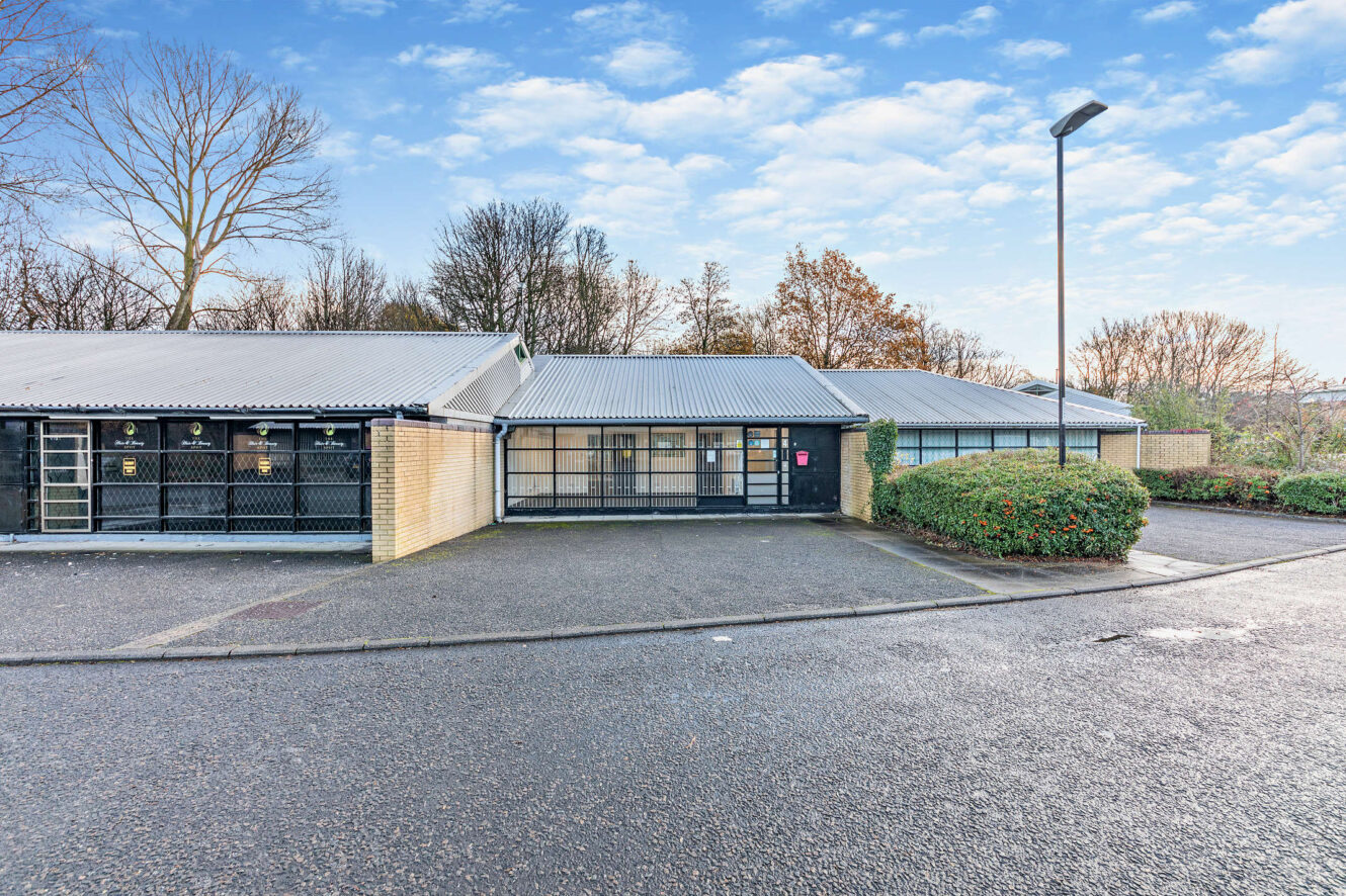Single-story commercial building with brick and metal exterior, glass entrance, adjacent parking area, and some bushes; trees are visible in the background under a partly cloudy sky.