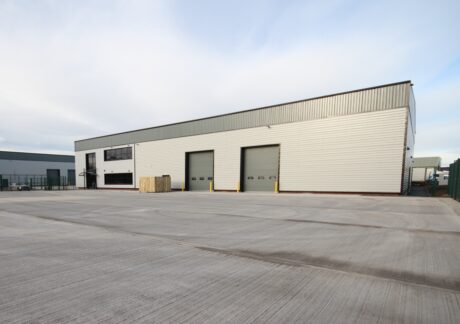 Large industrial warehouse with two gray loading bay doors, a small upper-level office section, and an expansive concrete lot in front.