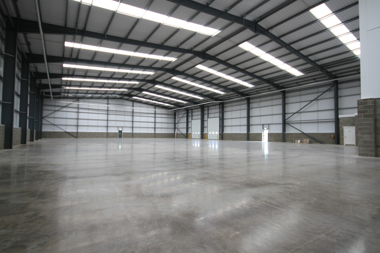 An empty, large warehouse with polished concrete floors, high ceilings, exposed steel beams, and several white doors along the walls.