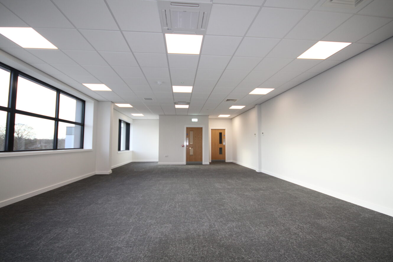 Empty modern office space with grey carpet, white walls, large windows on the left, and double wooden doors at the far end under a ceiling with recessed lighting.