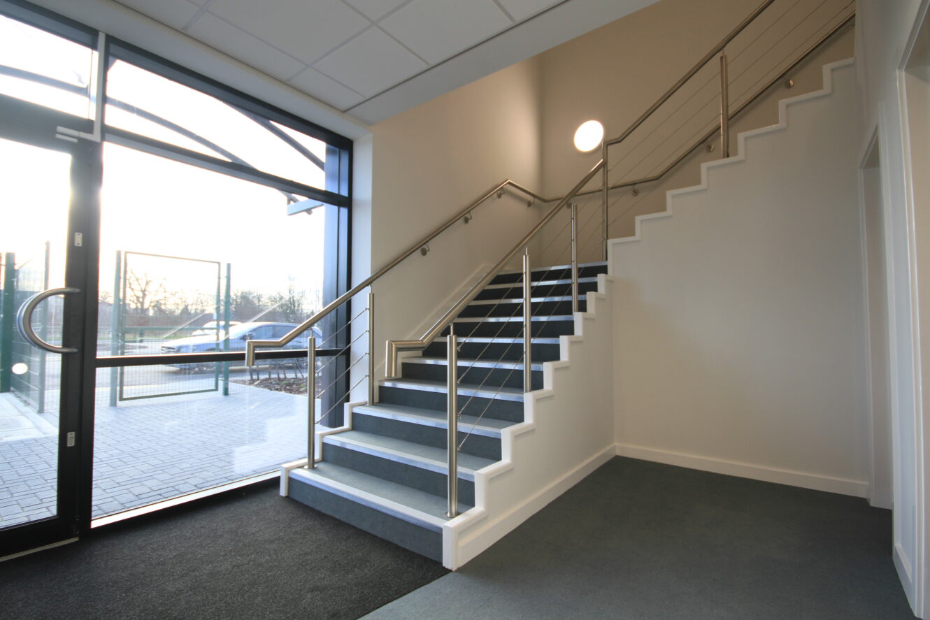 A modern indoor staircase with metal railings leads to an upper floor, next to large glass doors and windows letting in natural light.