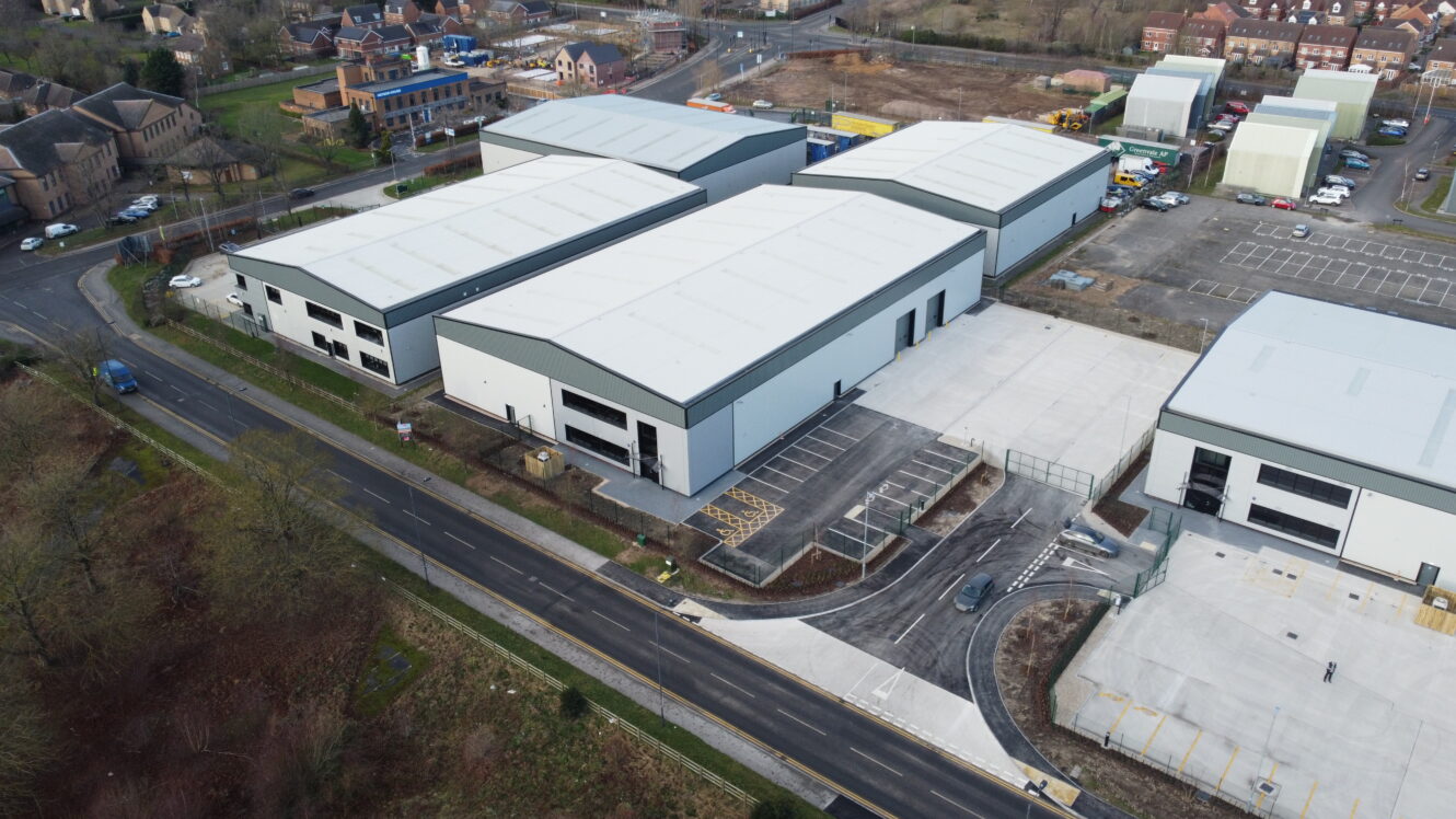 Aerial view of three large white industrial warehouse buildings with parking areas and access roads, surrounded by other structures and greenery.