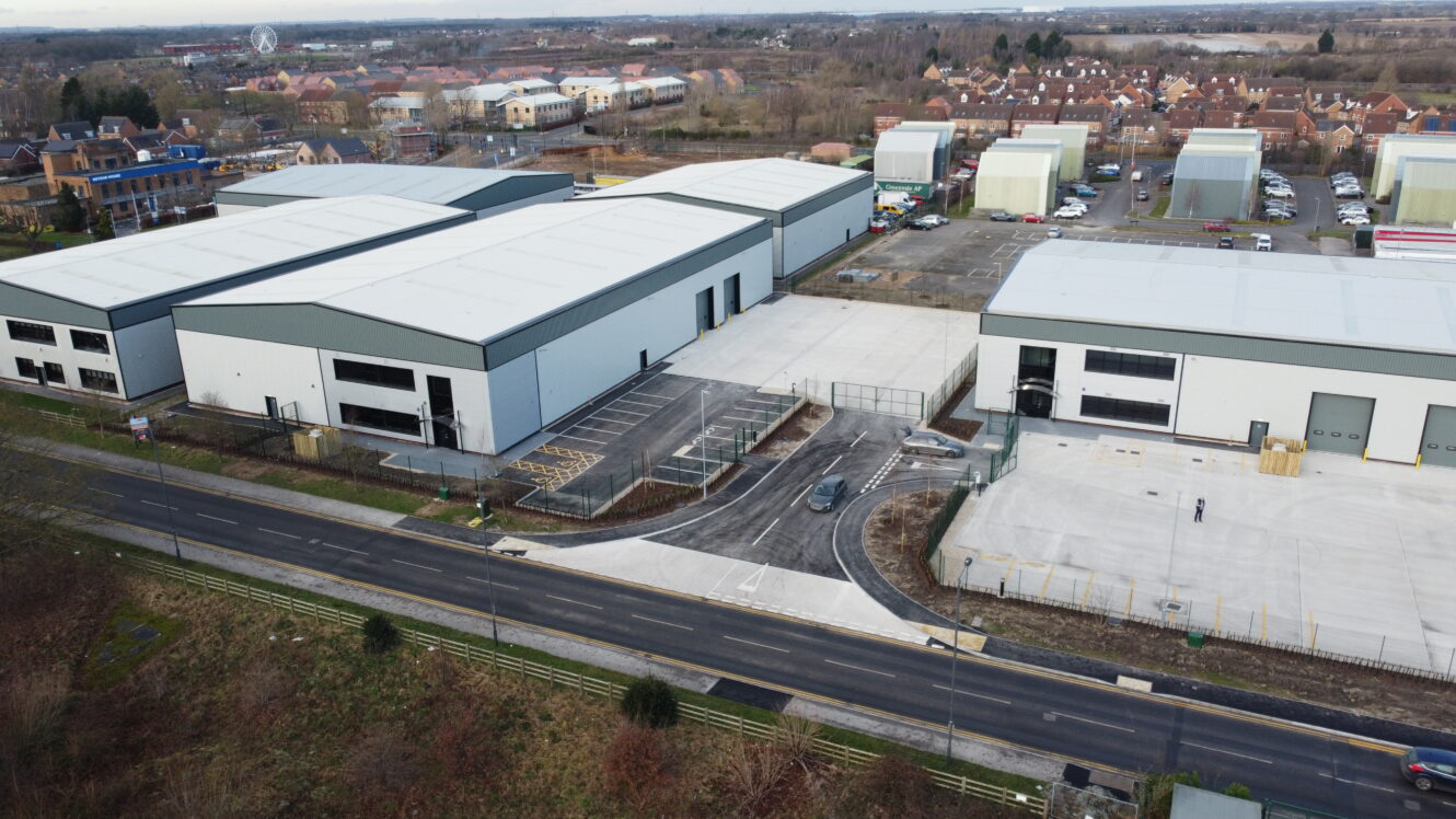 Aerial view of industrial warehouses with parking areas, surrounded by roads and residential houses in the background.