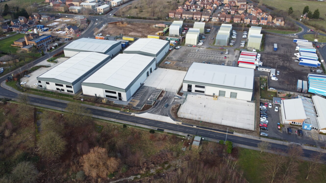 Aerial view of an industrial estate with multiple large warehouse buildings, parking lots, and a road running along the front, surrounded by residential areas and trees.