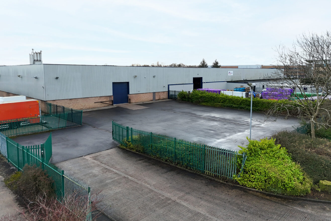 A large industrial warehouse with metal siding, fenced perimeter, and paved loading area; an orange container and stacks of purple-wrapped items are visible.
