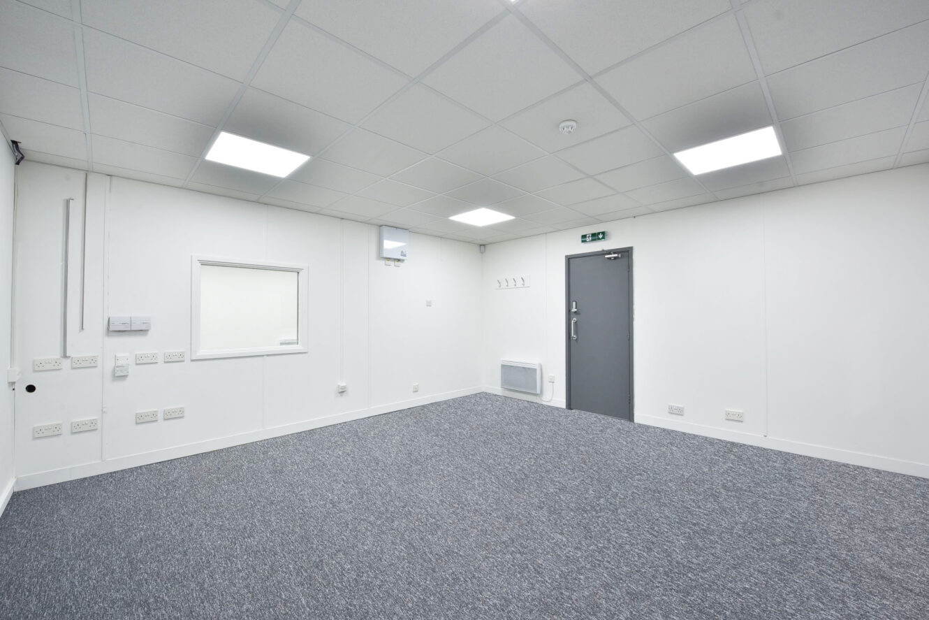 Empty modern office room with gray carpet, white walls, ceiling lights, a closed gray door, small window, and multiple electrical outlets.