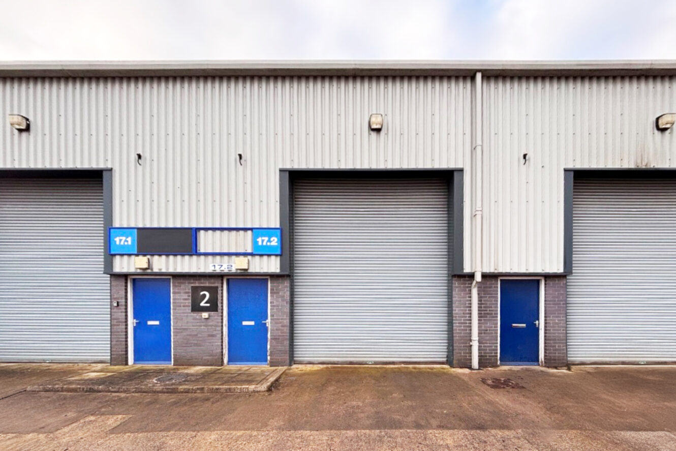 Industrial building exterior with three gray roller shutters and three blue doors, numbered “2,” “17.1,” and “17.2.” The ground is concrete and the wall is corrugated metal.