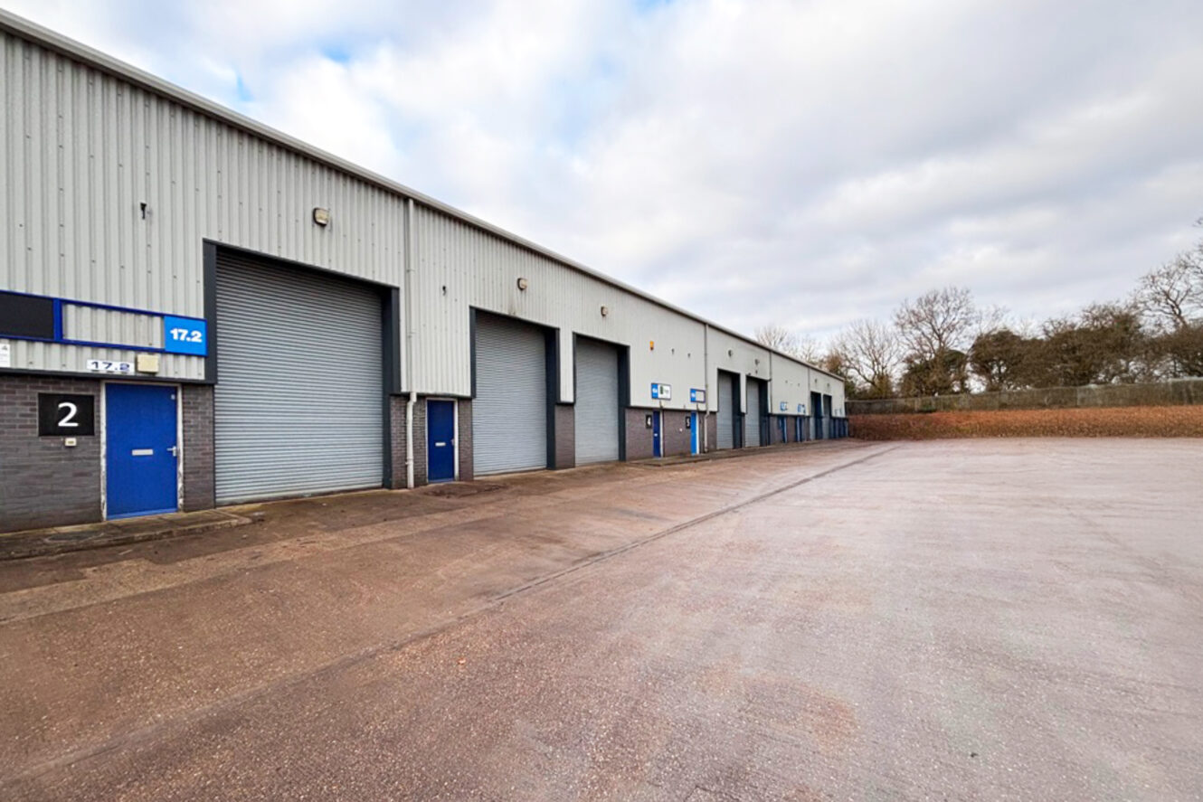 Row of industrial warehouse units with closed grey roller shutters and numbered doors, set along a spacious, empty paved yard under a cloudy sky.