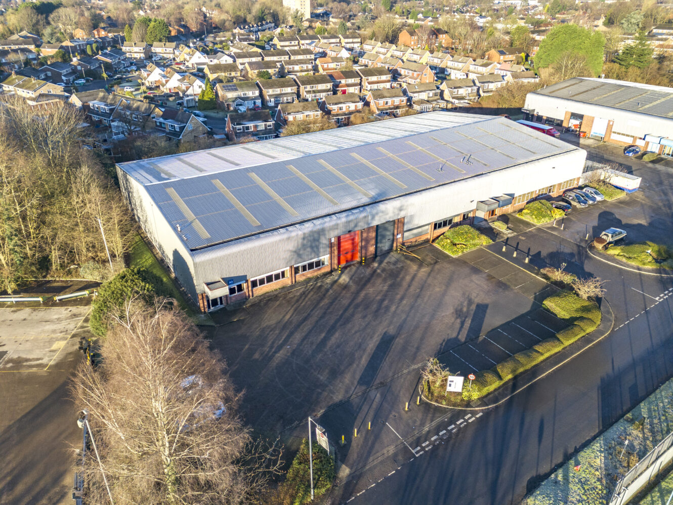 Aerial view of a large industrial warehouse with a metal roof, surrounded by a mostly empty parking lot and residential houses in the background.