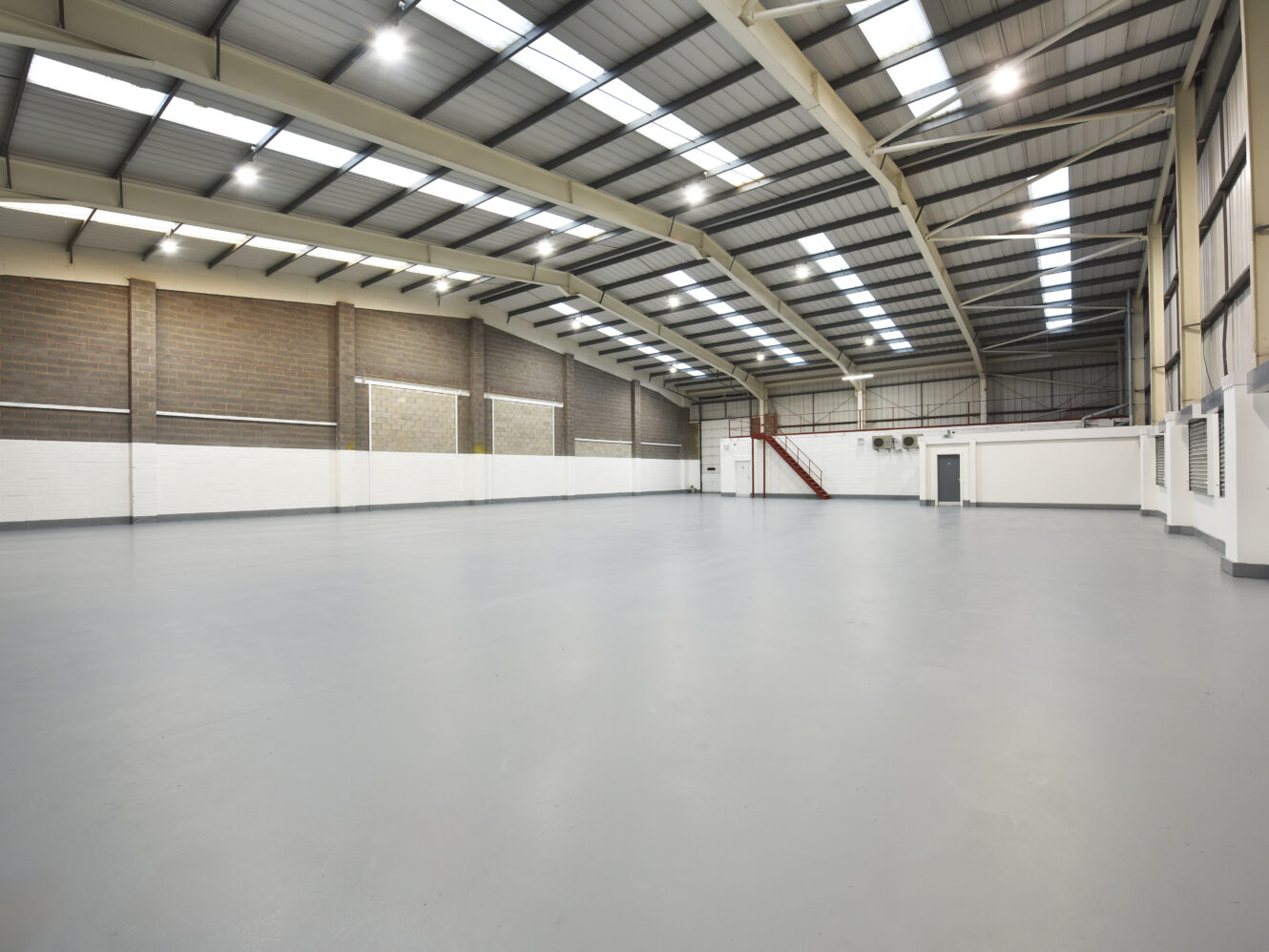 Large empty warehouse with high ceilings, concrete floor, brick and white walls, exposed beams, and a staircase leading to an upper area.