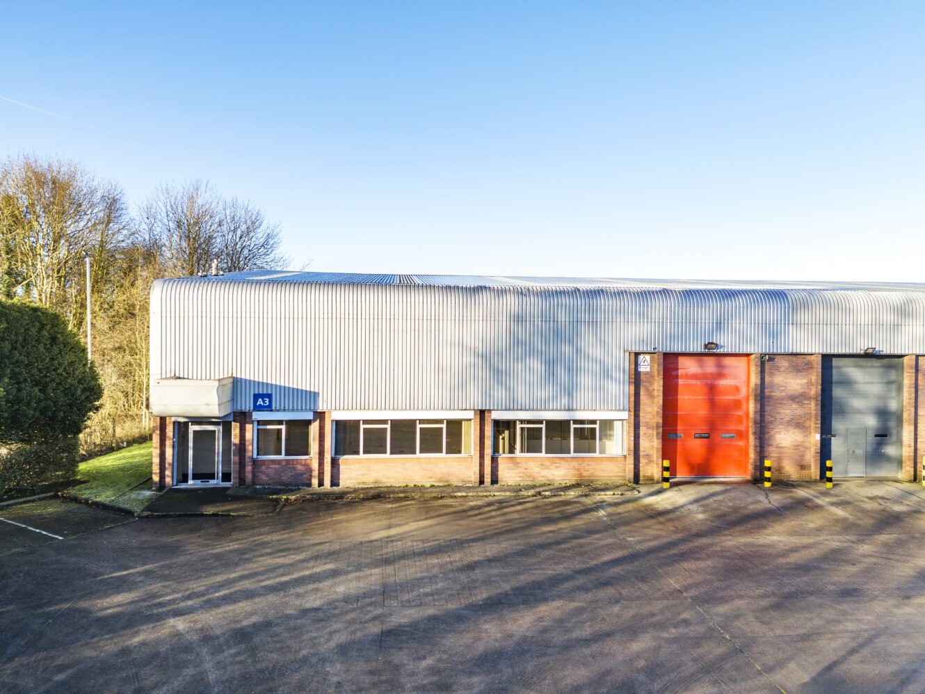 Industrial warehouse building with a corrugated metal roof, brick exterior, multiple windows, and large red and gray roller doors, set on a paved lot with trees in the background.