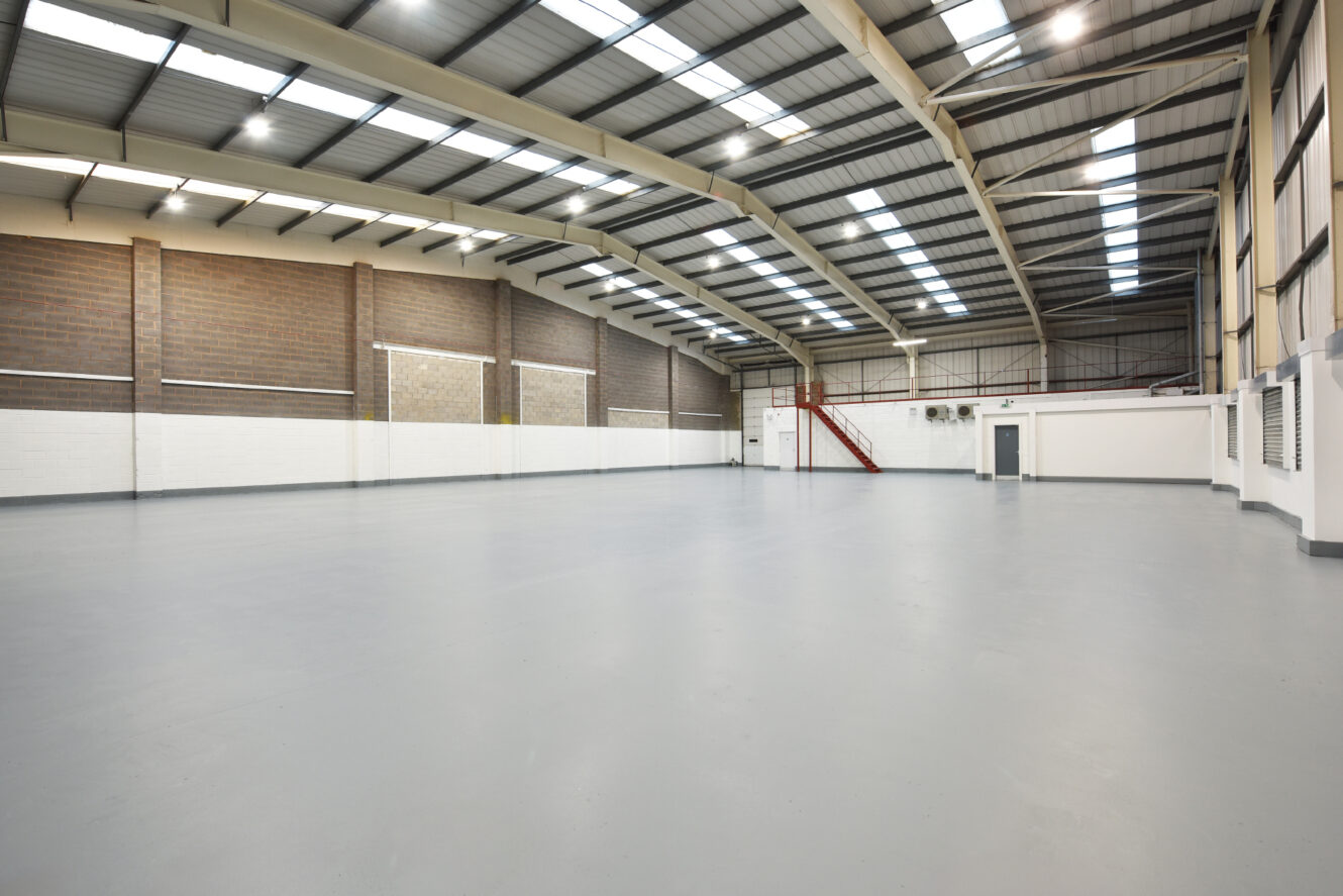 Large empty industrial warehouse with high ceilings, exposed beams, brick and white walls, and a red staircase leading to a mezzanine level.