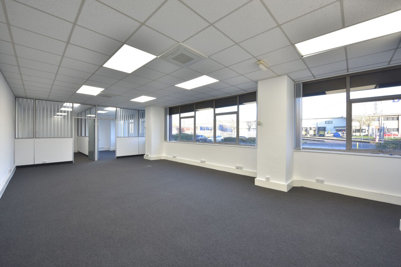 A spacious, empty office room with gray carpet, white walls, large windows, and fluorescent ceiling lights. Separate smaller office areas are visible in the background.