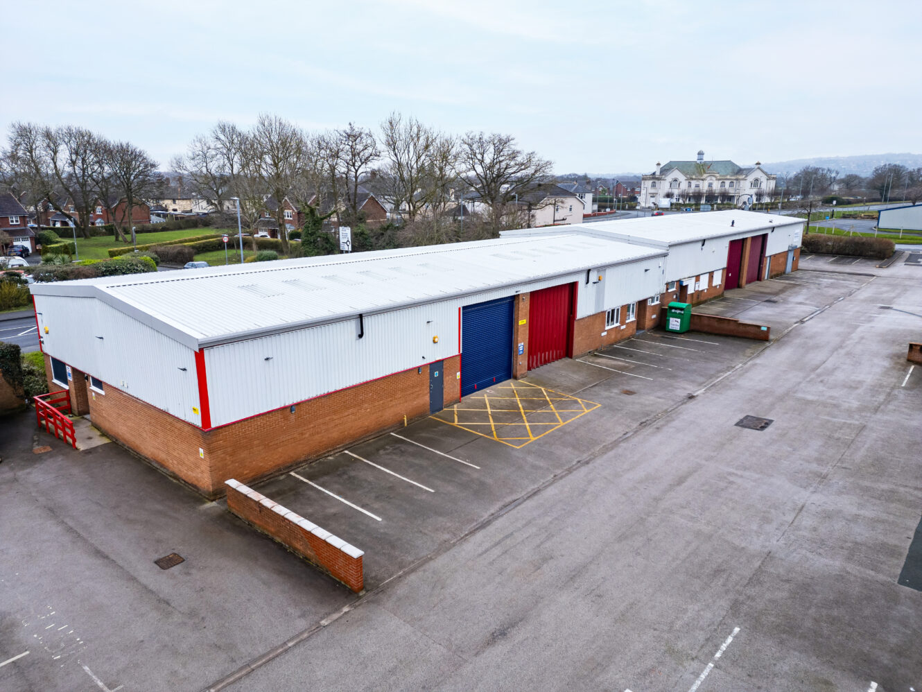 A row of industrial warehouse units with roller shutter doors, an empty parking lot, and a few trees and buildings in the background.