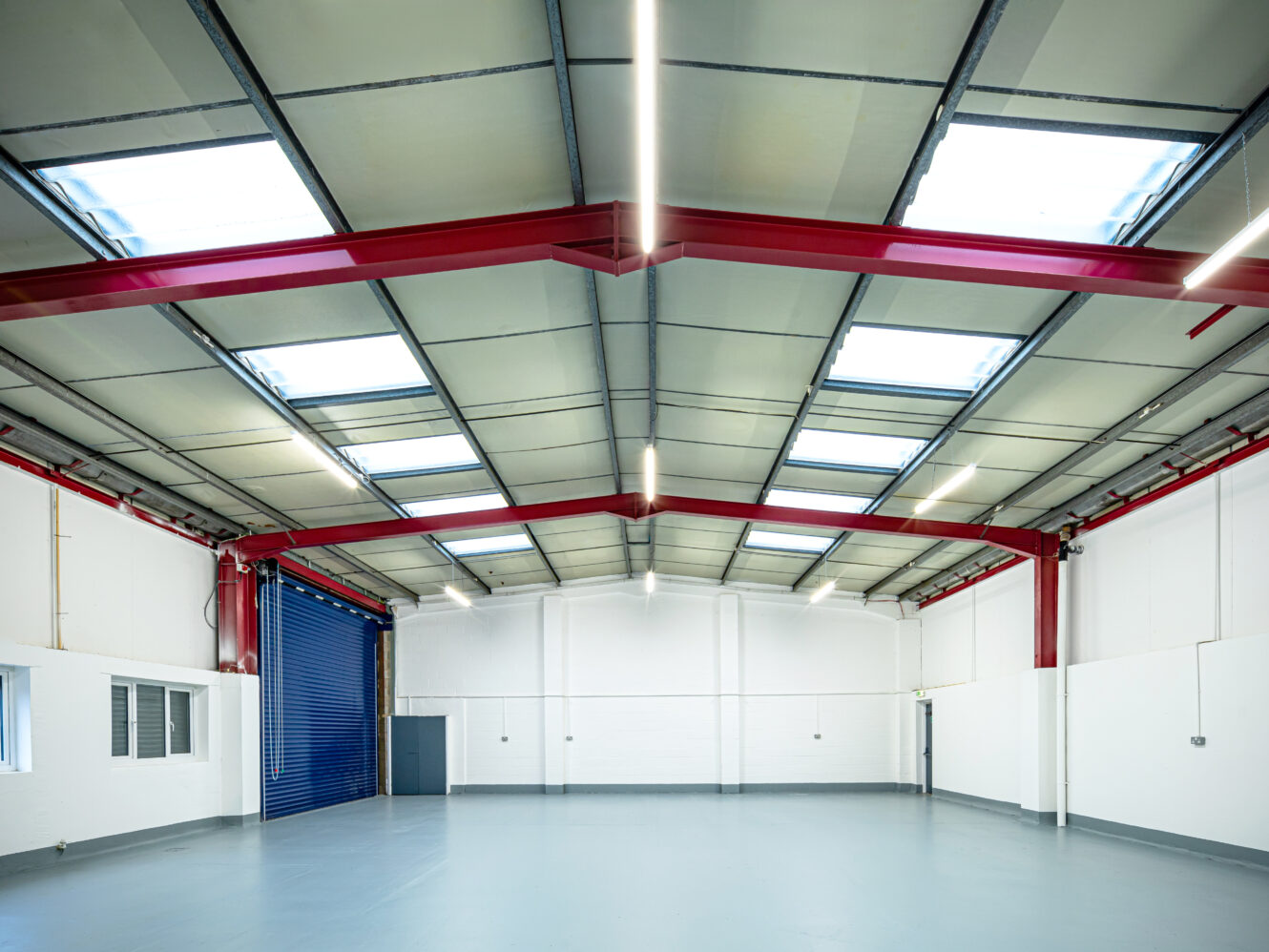 A clean, empty industrial warehouse interior with red steel beams, a high ceiling, skylights, white walls, and a large blue roller shutter door.