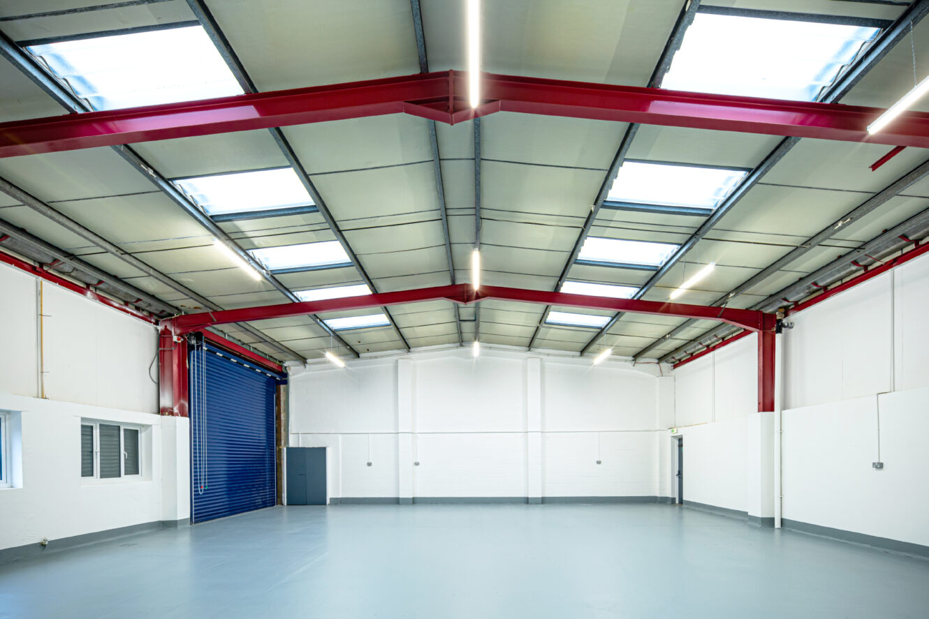 Empty industrial warehouse with gray flooring, white walls, red steel beams, skylights, and blue roller shutter door.