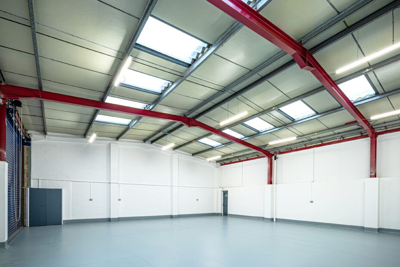 Interior of an empty industrial warehouse with a high ceiling, red steel beams, white walls, skylights, and a gray concrete floor.