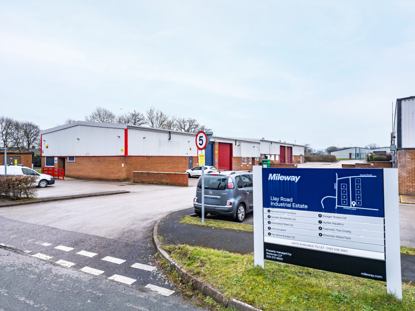 Entrance to an industrial estate with a directional sign, several parked cars, large warehouse buildings, and a 5 mph speed limit sign.