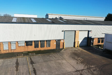 Large industrial warehouse with a brick lower section and metal upper walls, featuring a closed roller door and several boarded-up windows, set in an empty paved lot under a clear blue sky.