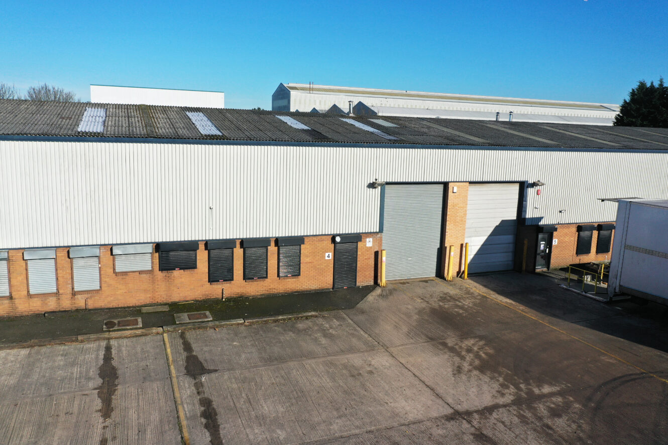 A large industrial warehouse with a corrugated metal roof, brick lower walls, closed shutters, and a tall roller door, set on a concrete lot under clear blue sky.