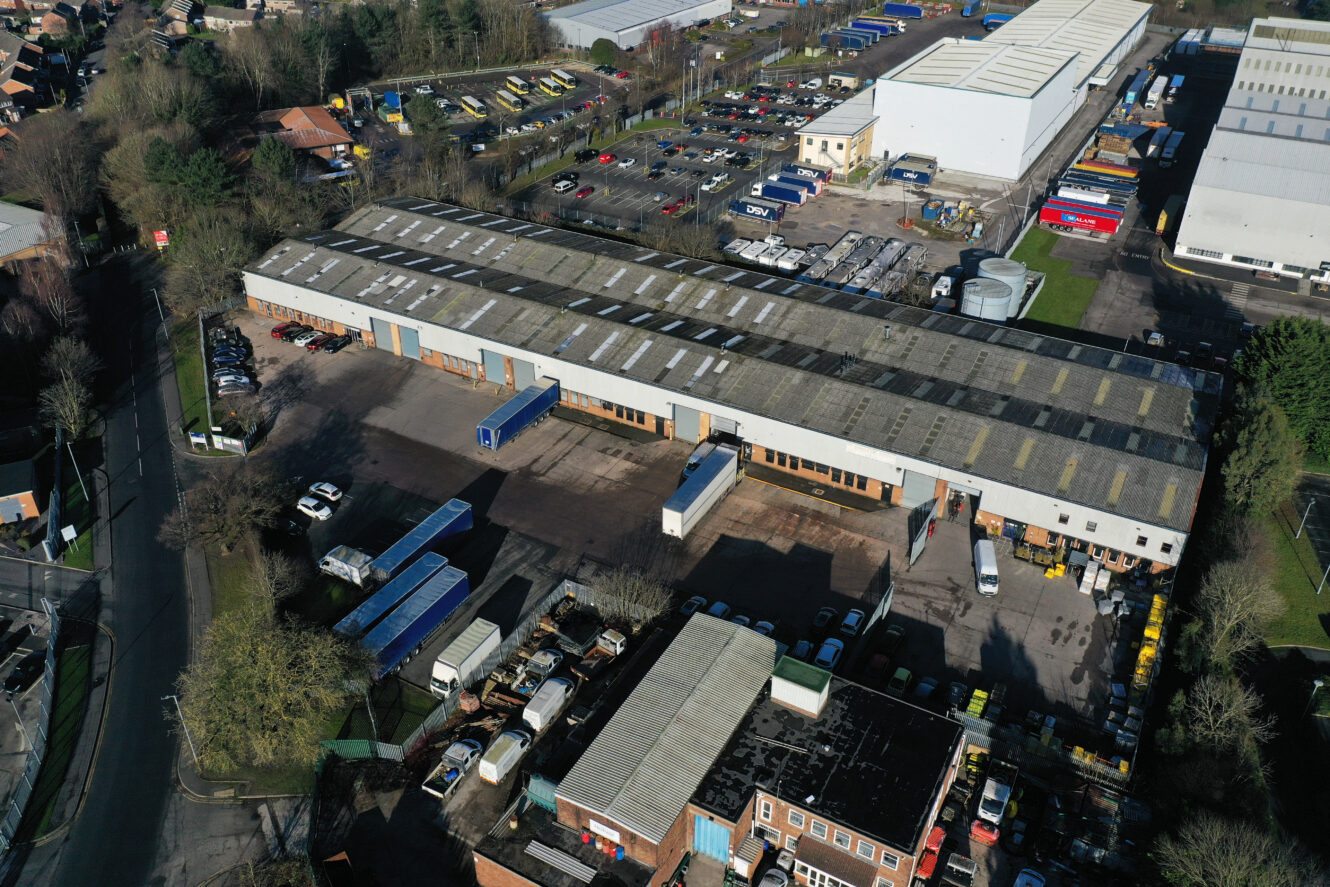 Aerial view of an industrial estate with warehouses, parked trucks, cars, and adjacent parking areas surrounded by trees and roads.