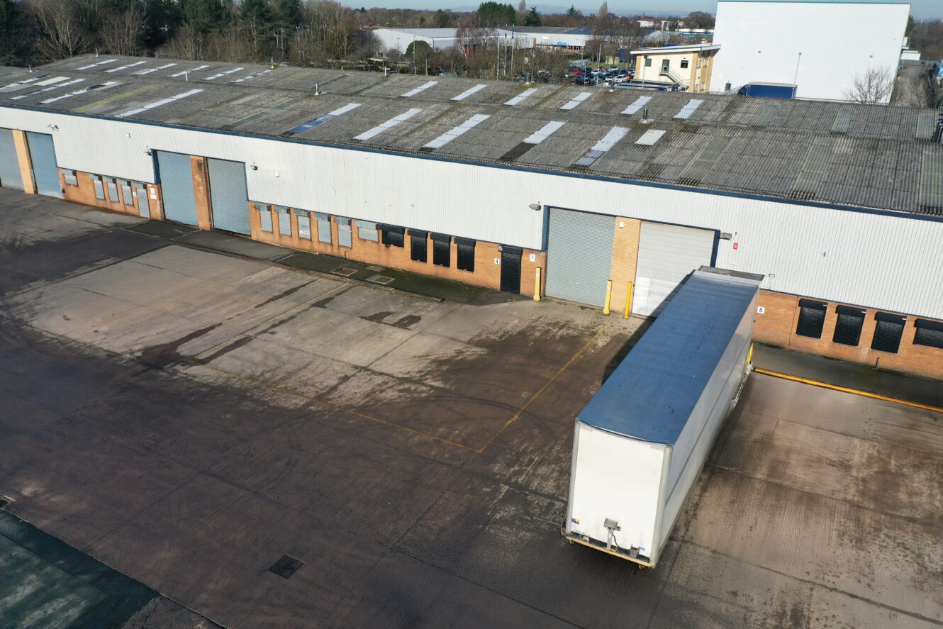 Aerial view of an industrial warehouse with a truck backed up to a loading bay on a large, mostly empty concrete lot.