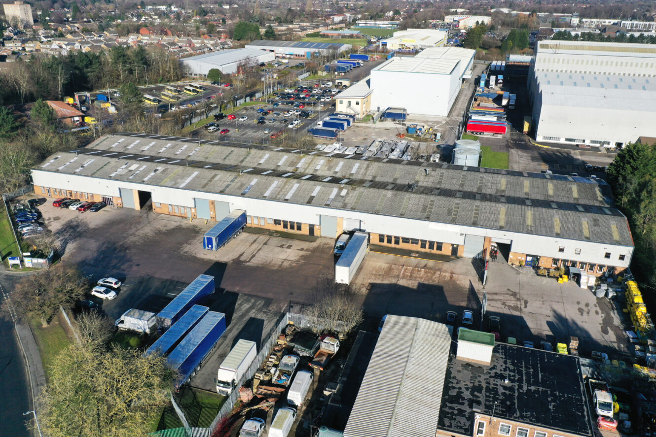 Aerial view of an industrial warehouse complex with parked trucks, cars, and adjacent buildings surrounded by trees and parking lots.