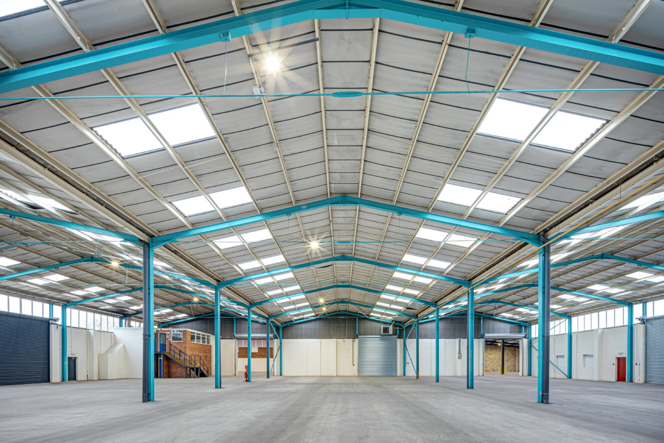 Interior of a large empty warehouse with a high ceiling, skylights, blue steel beams, and concrete floor.