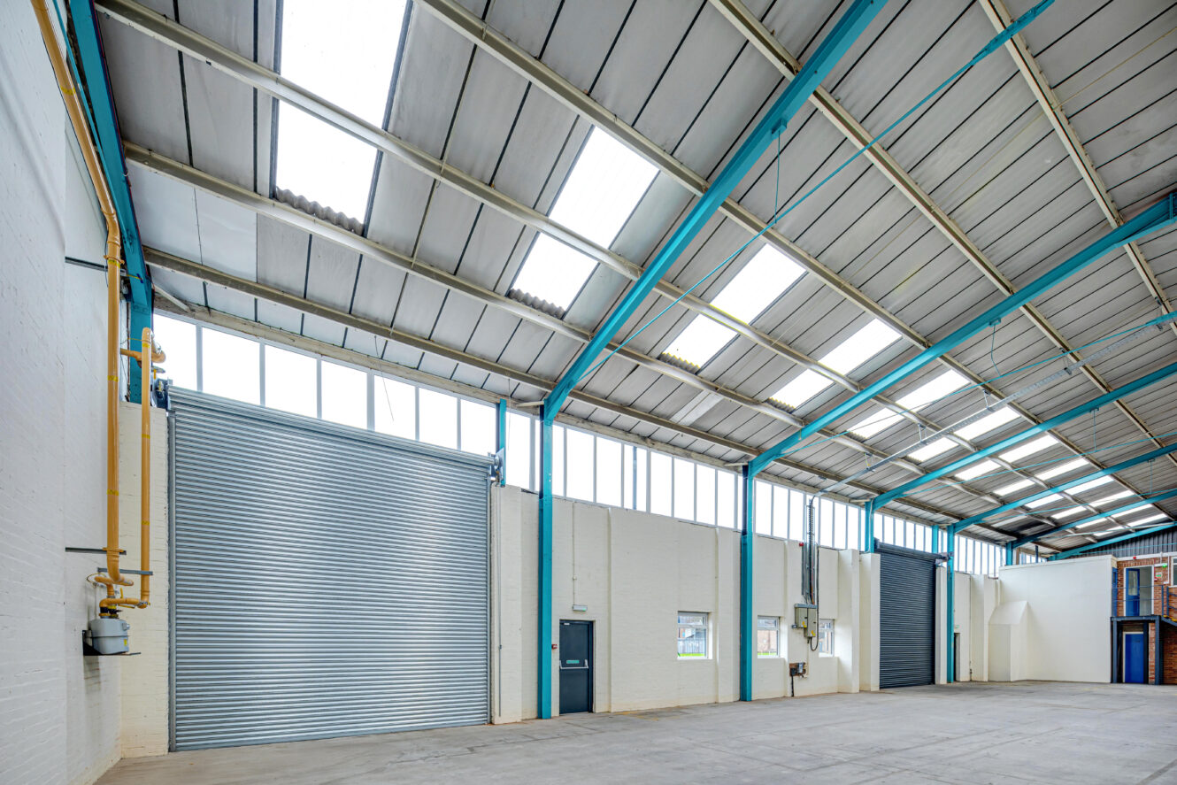 Spacious, empty warehouse interior with high ceilings, large windows, and two metal roller shutter doors.