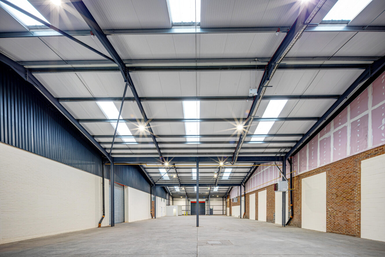 Interior view of an empty industrial warehouse with high ceilings, exposed beams, and overhead lights illuminating the space.