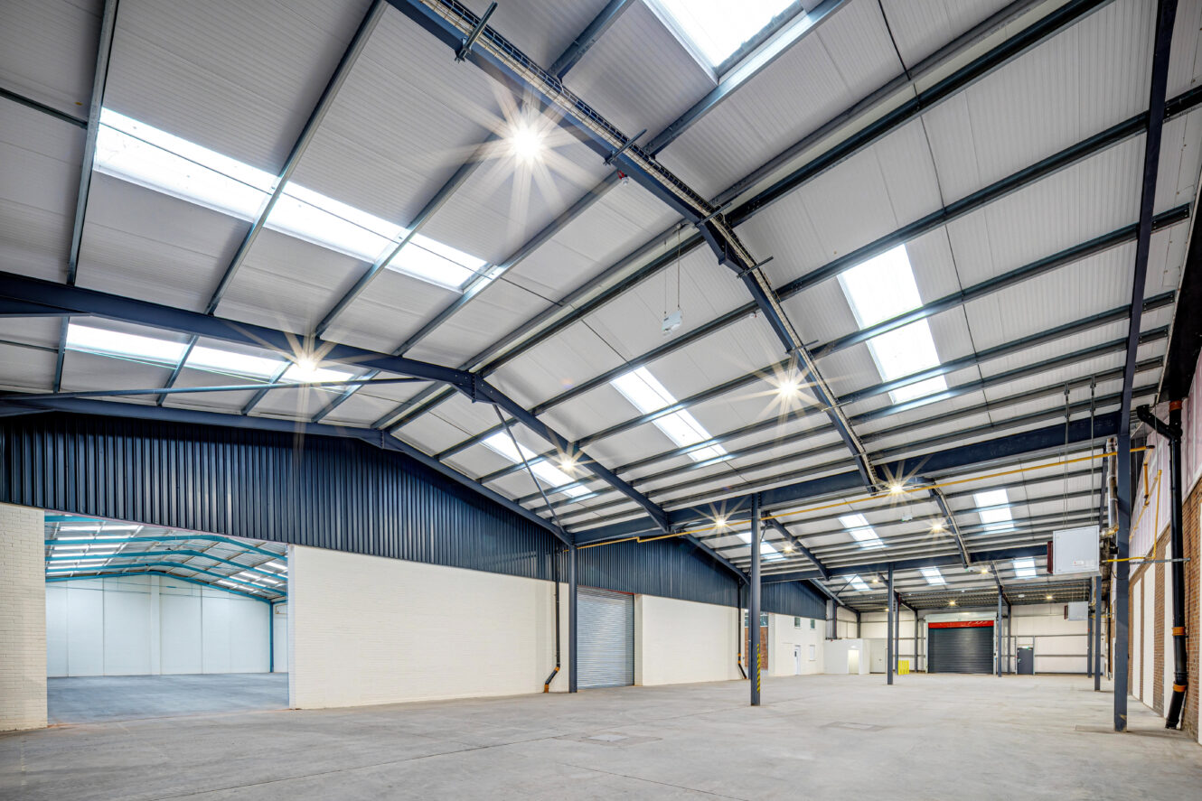 Spacious, empty warehouse interior with high ceilings, exposed steel beams, skylights, and large roller doors, illuminated by overhead lights.
