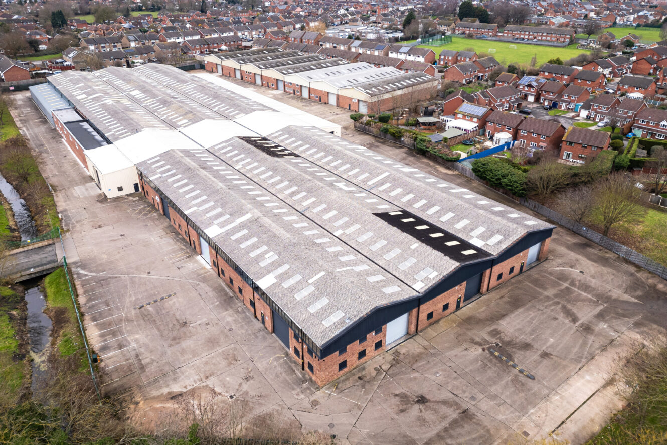 Aerial view of a large industrial warehouse complex surrounded by residential houses, with empty paved areas and some greenery nearby.