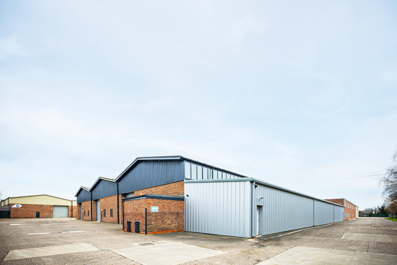 A large industrial warehouse with brick and metal siding, multiple garage doors, and an empty paved lot in front under a clear sky.