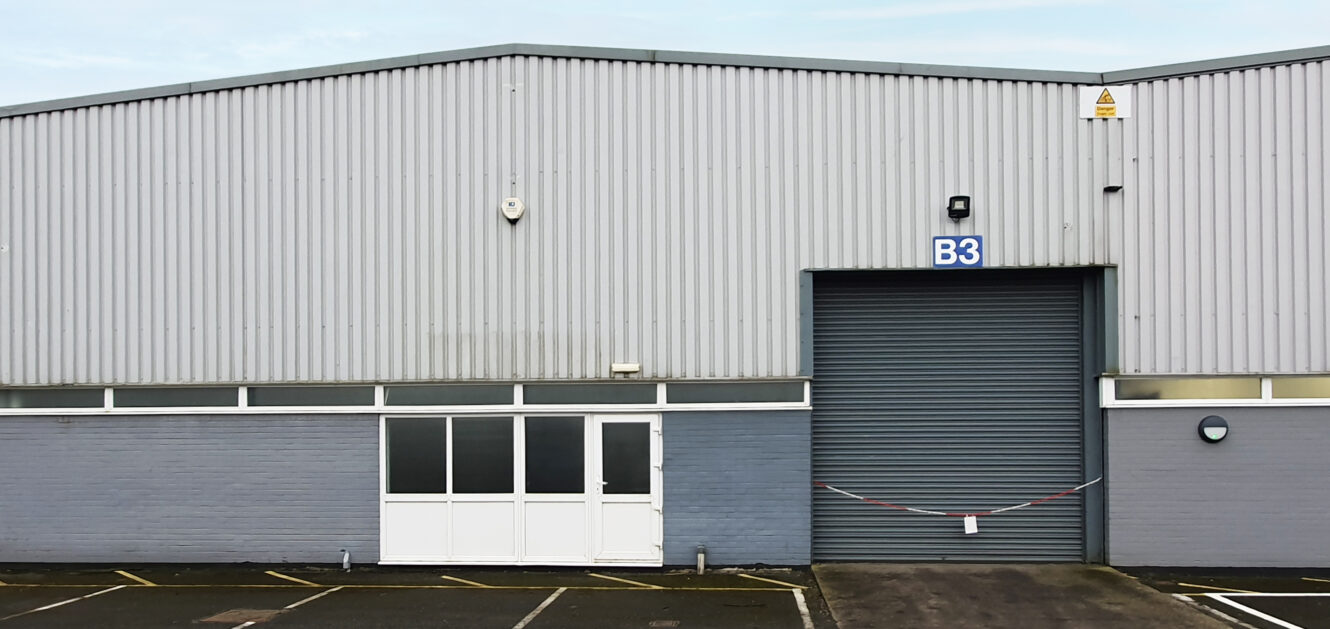 A large industrial warehouse with a gray metal exterior, a white entrance door, and a closed roller shutter labeled B3. A barrier tape is stretched across the shutter entrance.