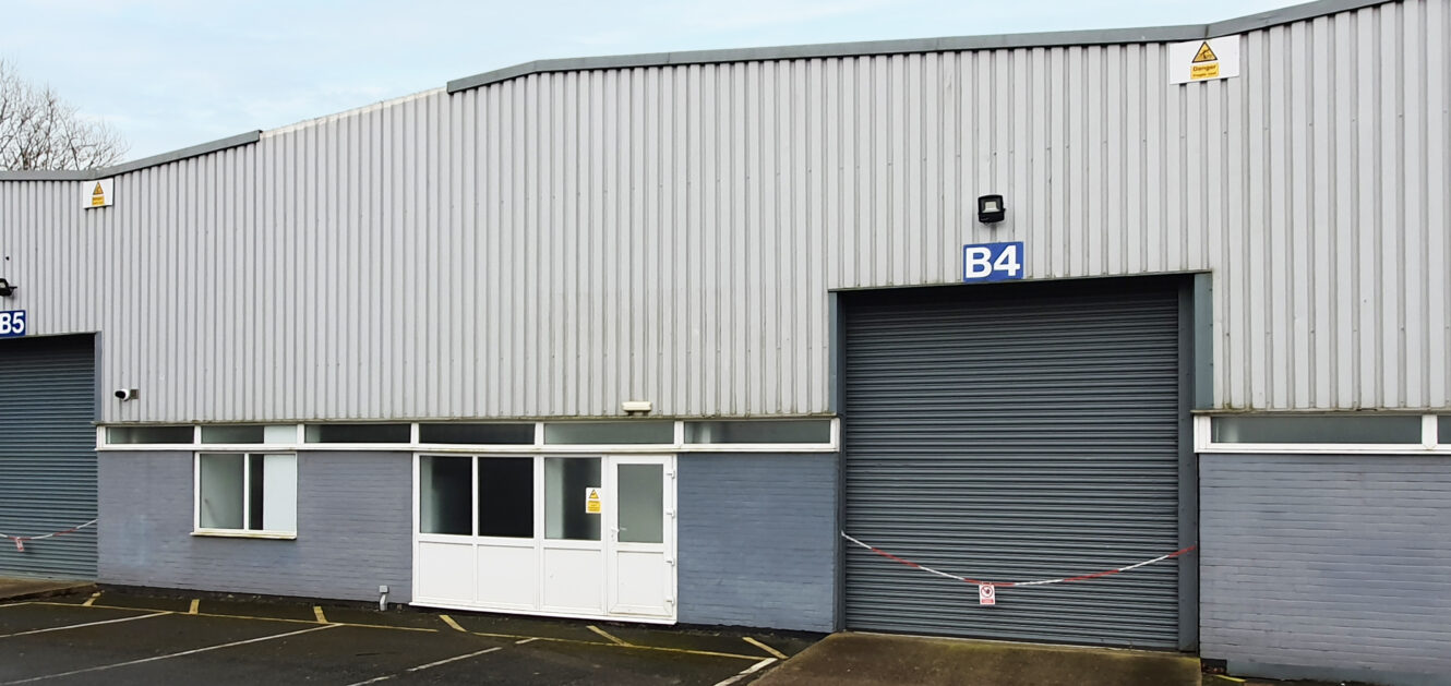 A grey industrial warehouse with a large roller shutter door labeled B4, adjacent to a white double door and parking spaces in front.