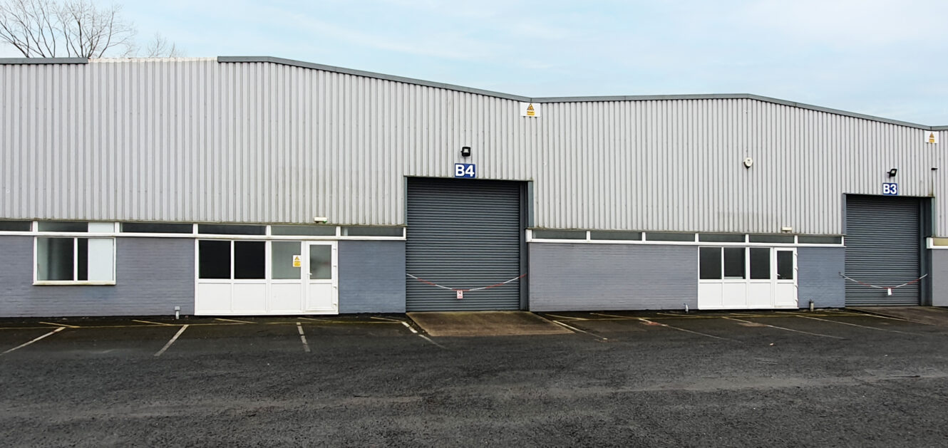 A gray industrial warehouse with unit labeled B4, a closed roller shutter door, white entry doors, windows, and an empty parking lot in front.
