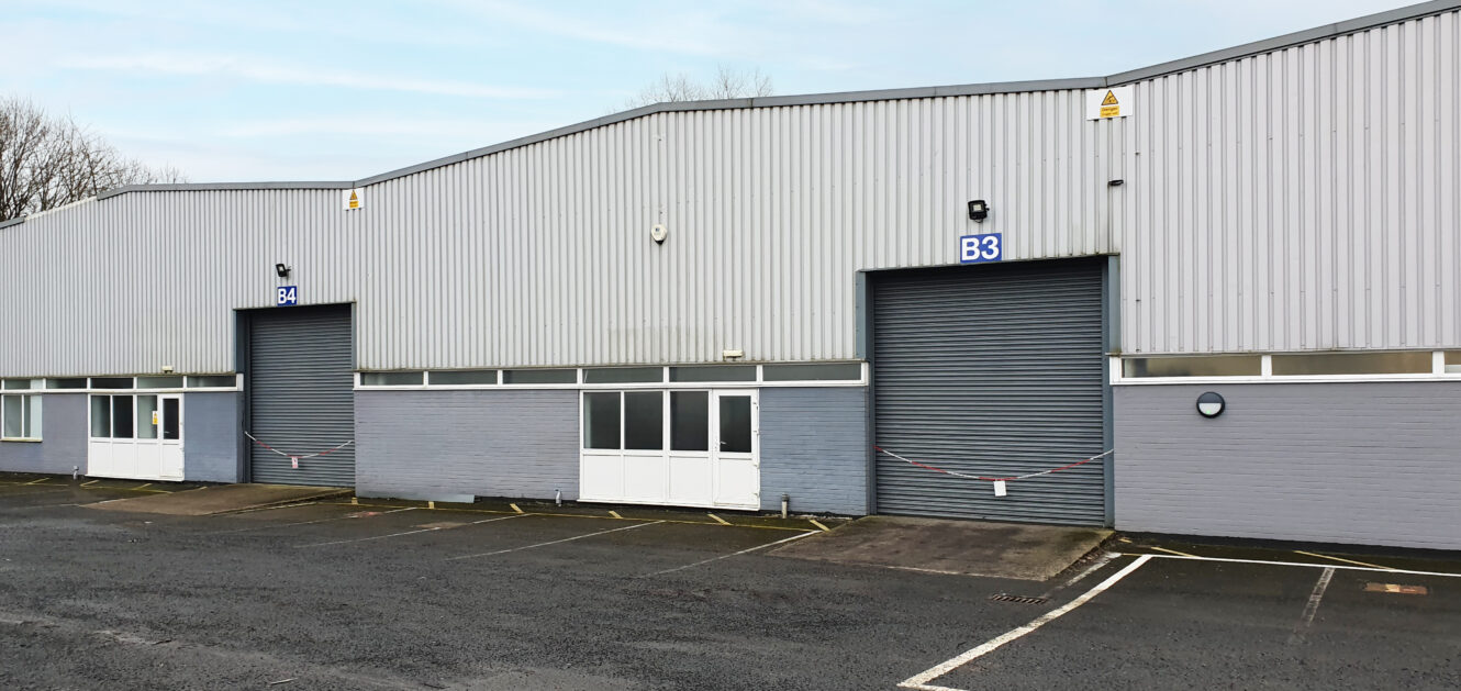A row of industrial warehouse units with grey roller shutter doors, labeled B3 and B4, and an empty parking lot in front.